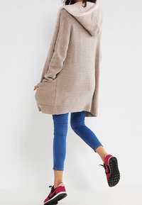 Beżowy, oversize'owy sweter z kapturem i puszystym wykończeniem, z dwoma bocznymi kieszeniami, w zestawieniu z niebieskimi rurkami i różowymi butami sportowymi.