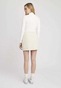Haut à manches longues blanc avec un col haut, associé à une mini-jupe en tweed jaune clair. Des baskets blanches et des chaussettes blanches visibles complètent l'ensemble.