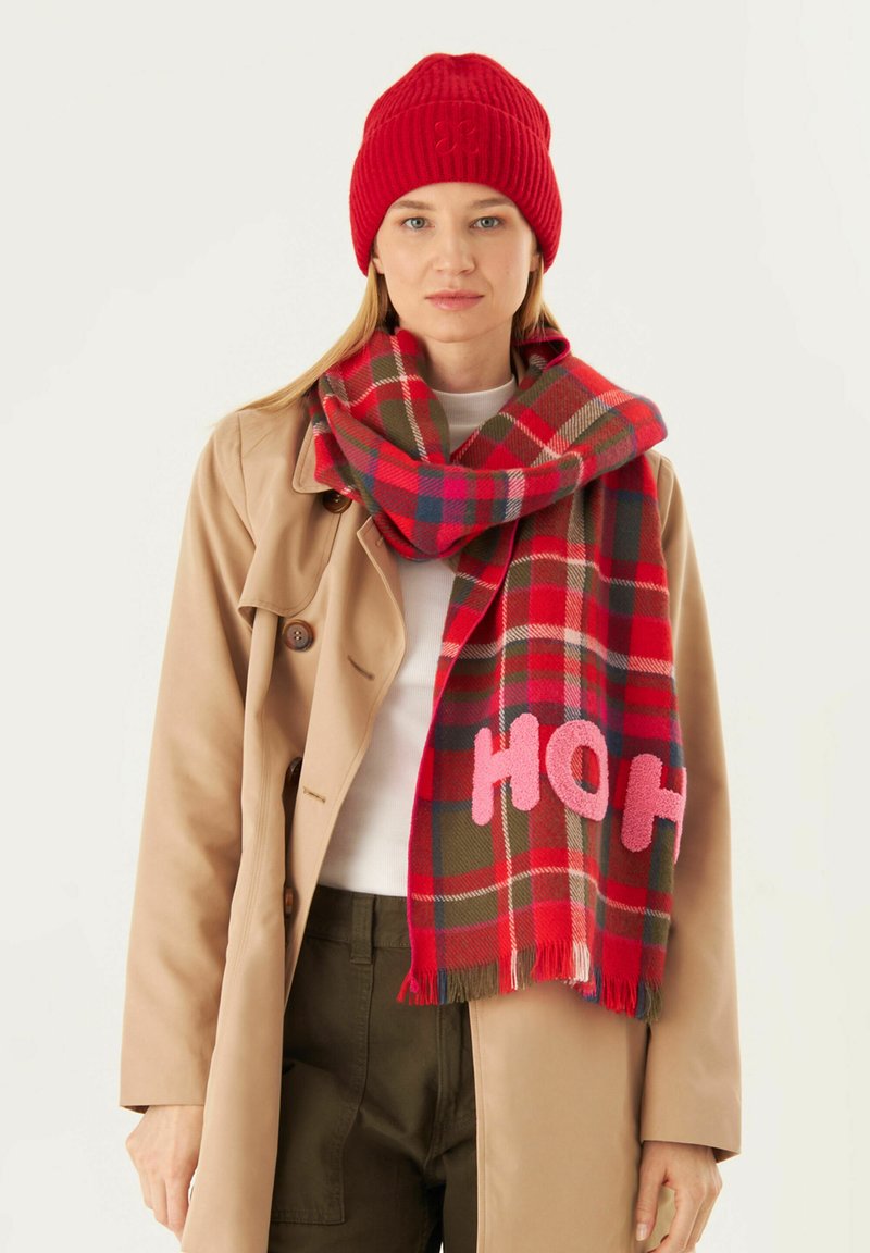 Rote ribbelige Mütze und karierschal in Rot, Grün und Blau, mit pinken "HOH" Buchstaben. Der Schal hat fransige Kanten und eine weiche Textur.