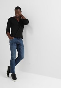 Zwarte lange mouwen henley shirt met knopen, gecombineerd met slim-fit blauwe denim jeans en zwarte sneakers, staande tegen een neutrale achtergrond.