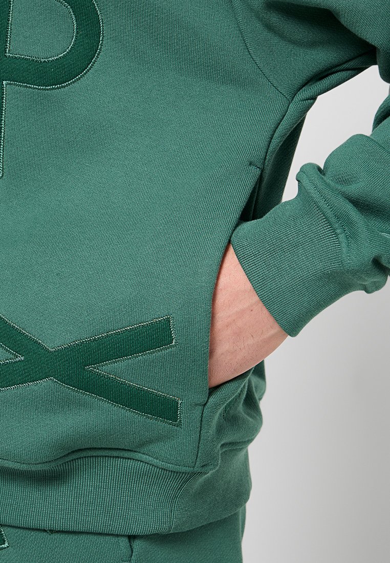 Sweatshirt vert avec une texture douce, présentant un grand "P" brodé sur le devant, des poignets côtelés et une poche sur le côté.