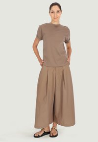 Brązowa bawełniana koszulka z podwiniętymi rękawami i brązowe szerokie spodnie z fałdami z przodu, w połączeniu z czarnymi sandałami.