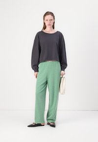 Szürke cropped pulóver, zöld textúrált nadrág és fekete lapos cipő. A modell egy bézs válltáskát tart, és egy fehér háttér előtt áll.