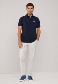 Polo shirt navy con colletto e logo ricamato, abbinato a pantaloni bianchi e sneakers blu, su uno sfondo neutro.