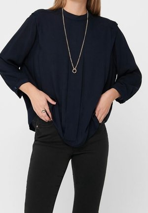 Vrouw draagt een loszittende donkerblauwe top met lange mouwen en zwarte broek, handen in de zakken, en een lange gouden ketting met een cirkelvormige hanger.