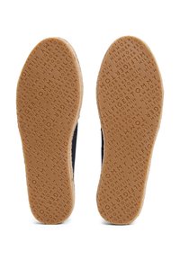Semelles de deux espadrilles bleu marine montrant des semelles en caoutchouc marron embossées d’un motif de texte répétitif « Tommy Hilfiger ».