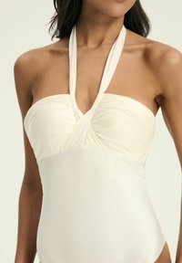 Kremowy jednoczęściowy strój kąpielowy z regulowanymi ramiączkami na szyję, marszczonym dekoltem i gładkim, elastycznym materiałem.