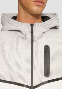 Grijze jack met rits van textuurstof, oversized capuchon en twee ritszakken. Kenmerkt zich door zwarte accenten en een logo op de rits.