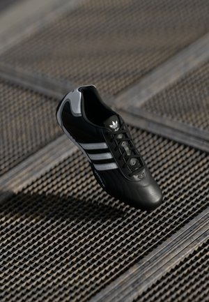 Black leather soccer shoe featuring three white stripes, textured surface, and a rounded toe. Accent details in gray on the heel area.