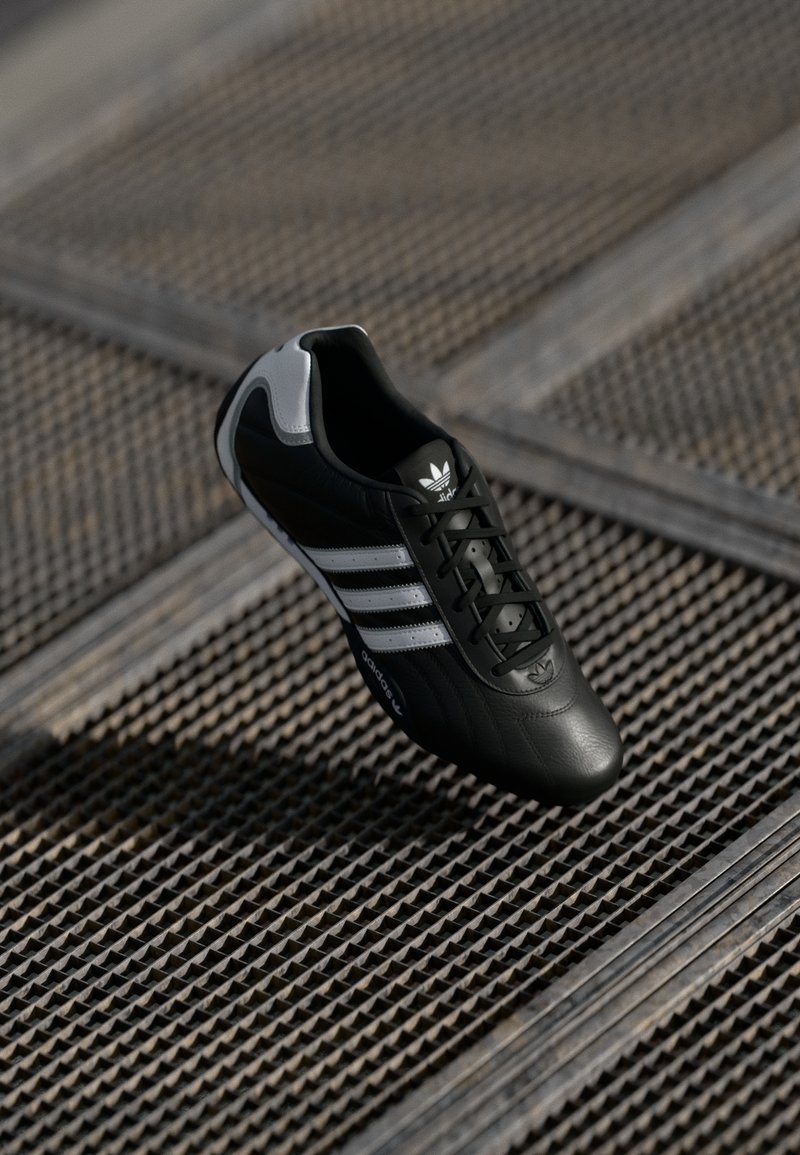 Black leather soccer shoe featuring three white stripes, textured surface, and a rounded toe. Accent details in gray on the heel area.