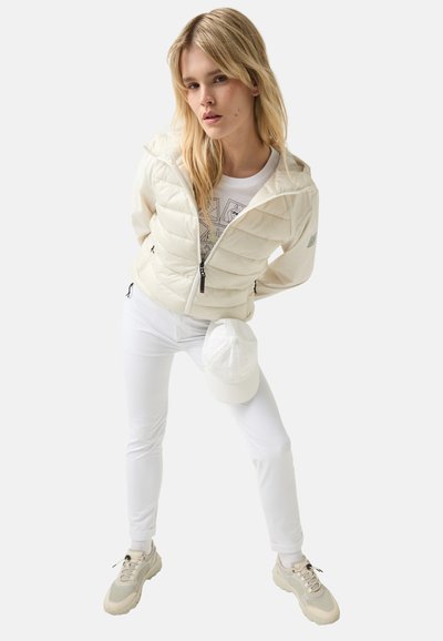 Hvid pufferjakke med lynlås foran, quiltet design og justerbar kant, parret med hvide leggings og snøresko. Hvid baseball-kasket på jorden.