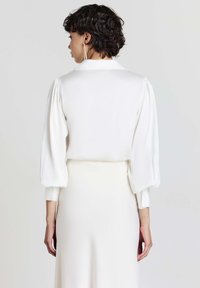 Blusa bianca con maniche a palloncino, morbido tessuto satinato e colletto, abbinata a una gonna liscia in un colore chiaro complementare.