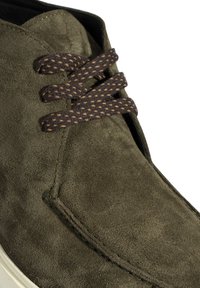 Groene suède schoen met een ronde neus, voorzien van bruine geweven veters en gestikte details langs de randen. Witte rubberen zool.