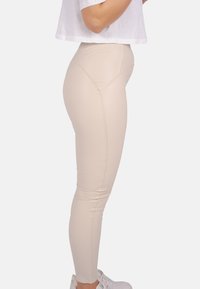 Beige Leggings aus elastischem Material mit taillierter Passform, versehen mit Nähdetails und hohem Bund, kombiniert mit weißen Sneakers.