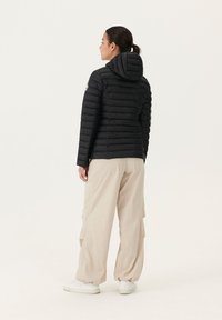 Zwarte pufferjack met horizontale stiksels en een capuchon, gecombineerd met losse beige broek en witte sneakers; gladde textuur en minimalistisch ontwerp.