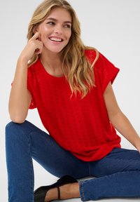 Haut rouge à manches courtes avec un motif en losange, associé à un jean slim bleu et des chaussures à talons noires. Texture lisse, décolleté rond.
