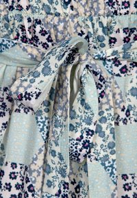 Blommig klänning med en blandning av blå, vit och marinblåa tyger. Har en rynkad byst och en knytning i midjan. Mönstren inkluderar små blommor och geometriska former.