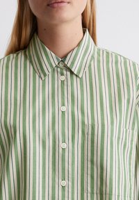 Camicia a righe verdi con strisce verticali in verde chiaro, bianco e rosso. Presenta un colletto, bottoni frontali e una tasca sul petto.
