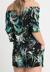 Kvinna klädd i en svart off-shoulder topp och matchande shorts med tryck av gröna, vita och blå tropiska blad och blommor.