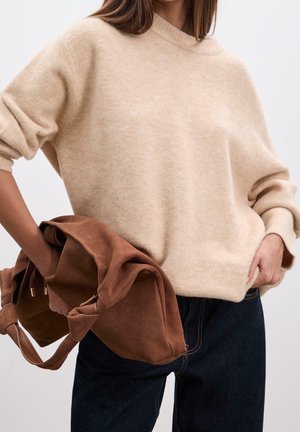 Femme portant un pull en laine beige duveteux et un jean foncé, tenant un sac à main en daim marron décontracté sur un fond clair uni.