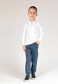 Biała koszulka henley z długim rękawem i trzema guzikami, zestawiona z ciemnoniebieskimi, zwężanymi spodniami. Dziecko nosi czarne trampki z białymi podeszwami.