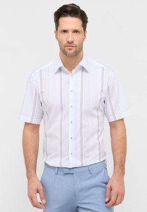 Mand iført en hvid kortærmet skjorte med tynde, lodrette striber og lyseblå bukser, stående foran en ensfarvet baggrund.