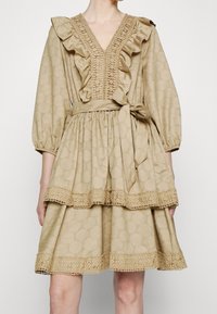 Robe beige avec un col en V, un col volanté et des manches ballon. Caractéristiques d'un tissu texturé, d'un design à étages et de détails en dentelle à l'ourlet.