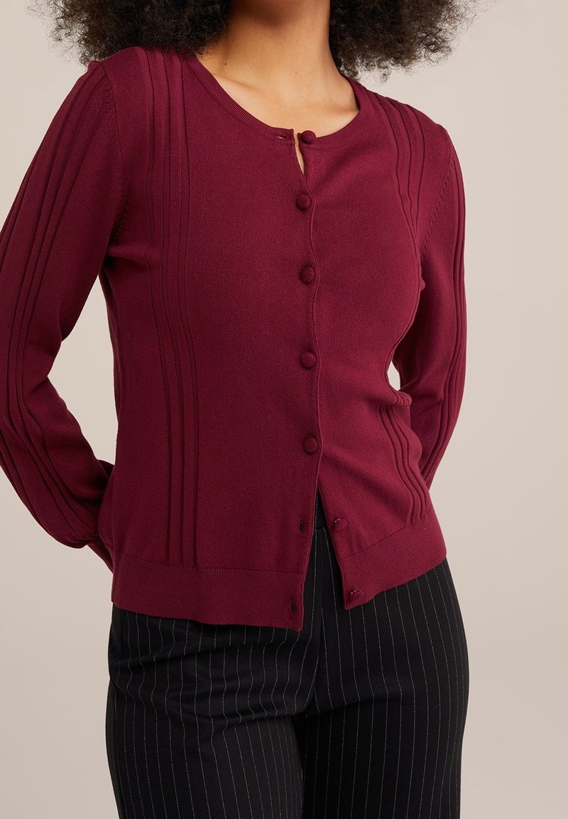 Vínově červený pletený kardigan s dlouhými rukávy, zapínáním na knoflíky a žebrovanými detaily podél ramínek. Kombinován s černými kalhotami s proužky.