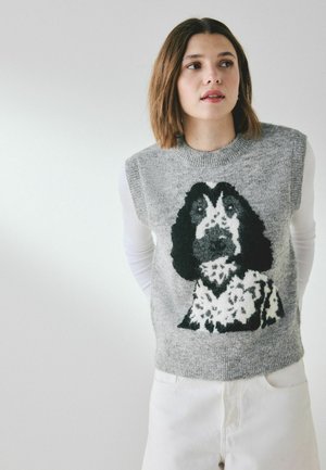 Frau mit schulterlangem braunem Haar trägt einen grauen ärmellosen Pullover mit einem schwarz-weißen Hunde-Motiv über einem weißen Langarmshirt und weißen Hosen.