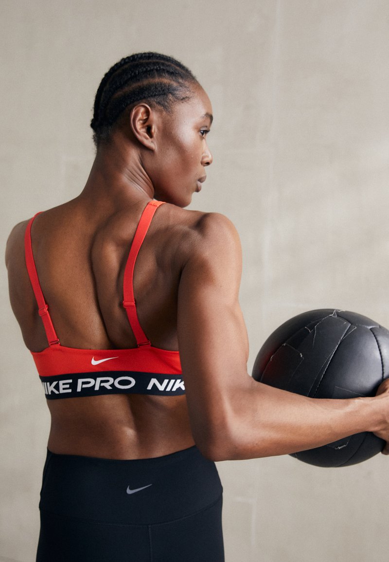 Sujetador deportivo rojo Nike Pro con banda negra, que presenta el logo. El modelo sostiene una pelota medicinal negra, mostrando músculos definidos y un físico atlético.