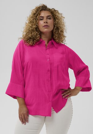 Femme aux cheveux bouclés portant une chemise boutonnée rose vif à manches larges et un pantalon blanc, posant avec une main sur la hanche devant un fond uni.