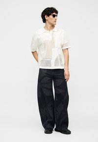 Homme portant des lunettes de soleil noires, un polo en maille blanc à manches courtes, un pantalon large noir à motifs et des chaussures noires, debout avec une main derrière le dos.