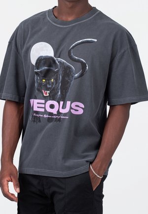 Osoba ubrana w ciemnoszarą koszulkę z wizerunkiem warczącej czarnej pantery, żółtymi oczami, pełnią księżyca oraz dużym fioletowym napisem "VEQUS".