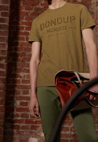 Olivgrön t-shirt med svart text, kombinerad med gröna byxor. Håller en ihopfälld beige väska med rödaktiga accenter och svarta handtag. Tegelvägg i bakgrunden.