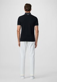 Camisa polo negra con cuello y puños de manga blancos en contraste, combinada con pantalones blancos y zapatos blancos. Figura de pie mostrada de espaldas.