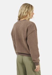 Donna con capelli biondi, che indossa un maglione marrone soffice e pantaloni verde oliva, vista da dietro mentre guarda di lato.