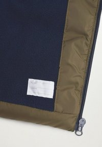 Granatowa polarowa kurtka wewnętrzna z oliwkowymi wykończeniami, widocznym zamkiem błyskawicznym i białą naszywką do wpisania imienia i numeru telefonu.