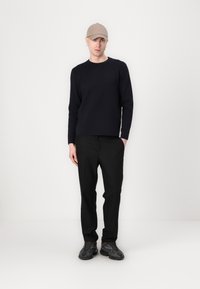 Marinefarbener Langarmpullover, schwarze Hose, beiger Hut und schwarze Schuhe mit strukturiertem Sohlen. Das Modell steht vor einem schlichten weißen Hintergrund.