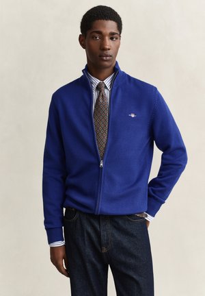 Jeune homme portant un pull bleu zippé par-dessus une chemise rayée et une cravate à motifs, debout avec une main dans la poche d’un jean foncé.