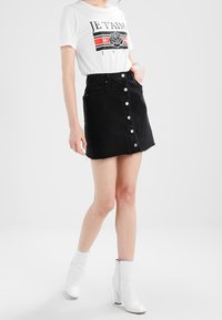 Falda de mezclilla negra con botones frontales, dobladillo deshilachado, combinada con una camiseta gráfica blanca y botas blancas hasta el tobillo. Textura suave, ajuste moderno.