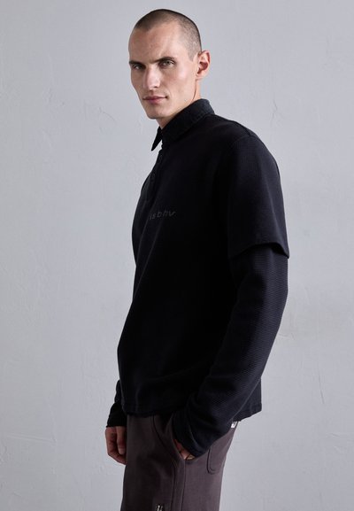 Mann mit kurzen Haaren, der ein schwarzes Langarmshirt aus Waffelstrick unter einem kurzärmligen schwarzen Hemd trägt und mit den Händen in den Hosentaschen steht.