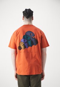 Hombre con cabello trenzado que lleva una camiseta naranja con un gráfico espiral abstracto colorido en la parte posterior, de pie contra un fondo blanco liso.