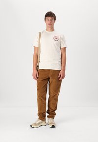 Beige t-shirt med rød grafik, brune kordfløjspants og beige sneakers med lyse accenter, stående mod en hvid baggrund.