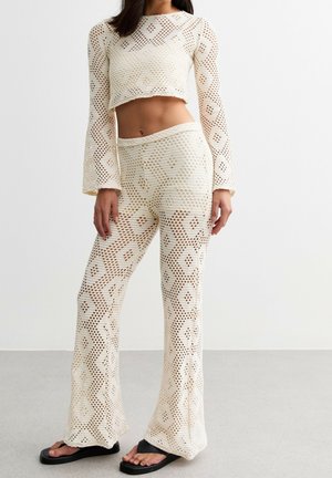 Mujer con top corto de manga larga de crochet color crema y pantalones acampanados a juego con estampados florales, combinados con chanclas negras.