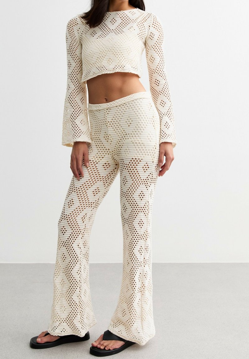 Femme portant un crop top en crochet crème à manches longues et un pantalon évasé assorti à motifs floraux, associé à des tongs noires.