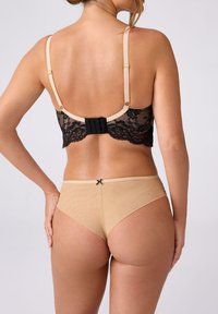 Kobieta nosząca beżowe siatkowe majtki z czarną kokardą oraz czarny stanik koronkowy z beżowymi ramiączkami, pokazana od tyłu na jednolitym tle.