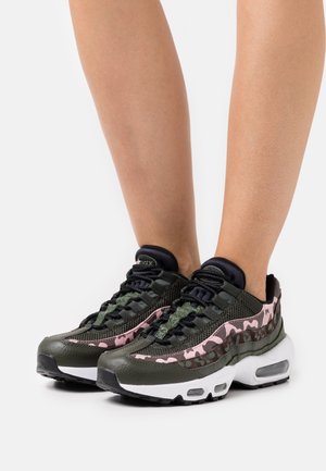 Jambes chaussées de baskets noires et verts olive avec des touches de camouflage rose et des semelles blanches, debout sur un fond blanc.