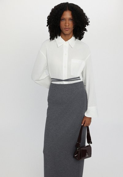 Camisa blanca de botones con un bolsillo, combinada con una falda gris ajustada. En la mano derecha se sostiene un pequeño bolso burdeos con un acabado brillante.