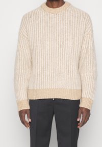 Beige gestrickter Pullover mit geripptem Muster, Rundhalsausschnitt und lockerer Passform. Lange Ärmel mit gerippten Bündchen; der Saum ist ebenfalls gerippt.