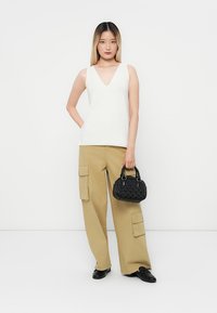 White v-neck tank top, beige cargo pants with large pockets, and a textured black handbag. Model stands against a plain white background.